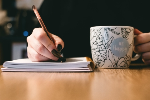 A person writing in a notebook and holding a mug.