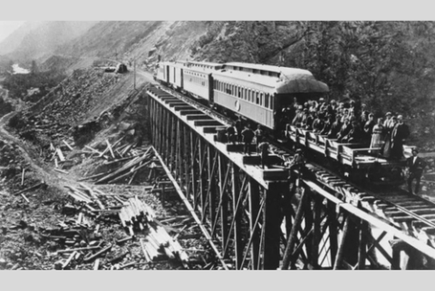black-and-white photo of five train cars, with a flatbed car at the front carrying people on a train track next to a mountain side.