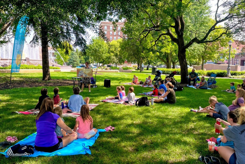 Librarian reading a book in the park with families watching from blankets.