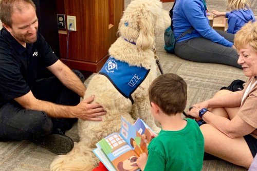 Children reading to a dog and the dog's owner.