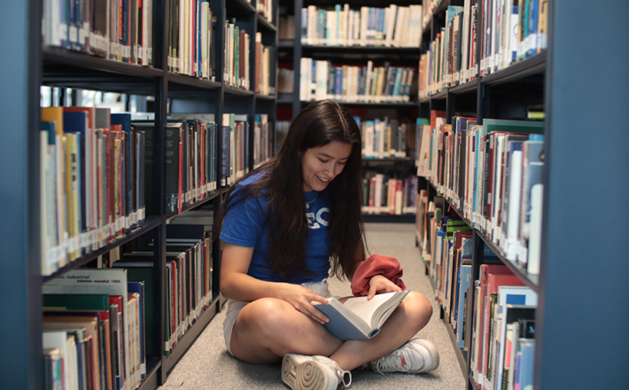 Teen girl sitting between book stacks with open book in lap